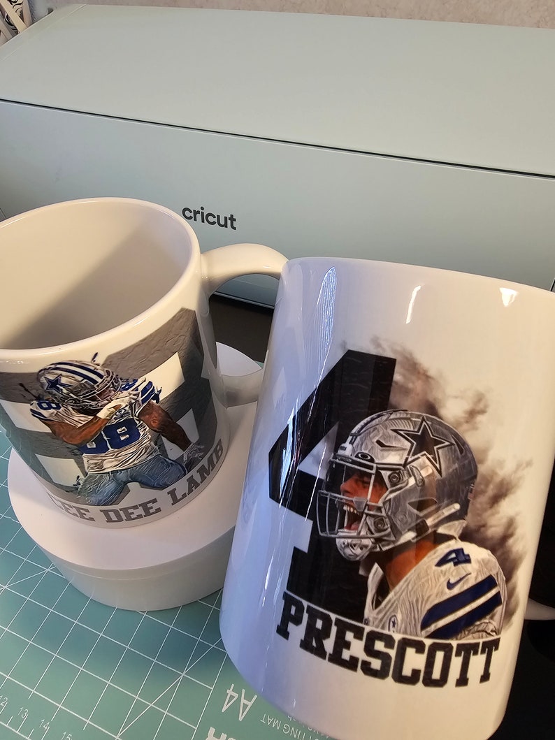 May include: Two white ceramic mugs with images of Dallas Cowboys football players. One mug features a player in a blue jersey with the number 88 and the name "Cee Dee Lamb". The other mug features a player in a blue jersey with the number 4 and the name "Prescott".