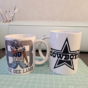 May include: Two white ceramic mugs with a Dallas Cowboys theme. One mug features a blue and white image of CeeDee Lamb, a Dallas Cowboys wide receiver, with the number 88. The other mug features a large blue star with the word "COWBOYS" in black lettering.