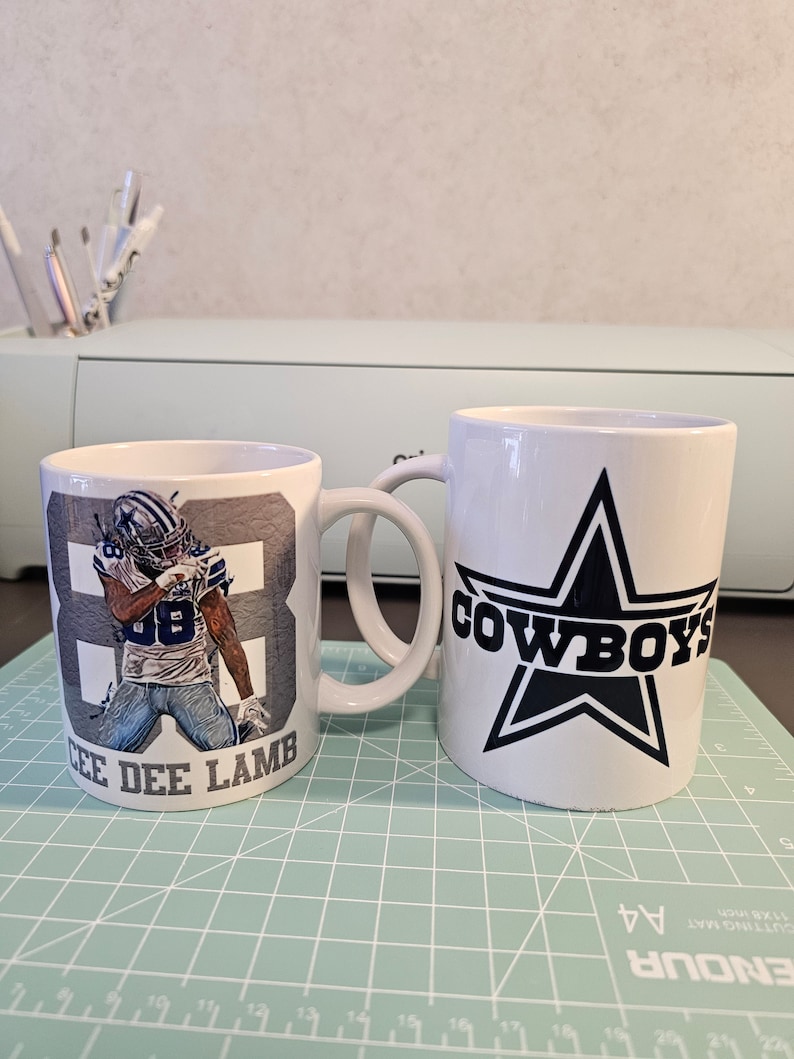 May include: Two white ceramic mugs with a Dallas Cowboys theme. One mug features a blue and white image of CeeDee Lamb, a Dallas Cowboys wide receiver, with the number 88. The other mug features a large blue star with the word "COWBOYS" in black lettering.