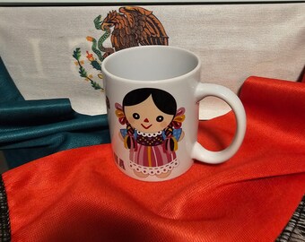 May include: White ceramic mug with a colorful illustration of a traditional Mexican doll. The doll is wearing a pink, red, and white dress with black hair and a red bow. The mug has a black handle and the text "Viva Mexico" is printed on the front.