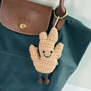 Op de afbeelding: Een gehaakte bruine cactus sleutelhanger met een glimlachend gezicht en twee kleine bruine benen. De sleutelhanger is bevestigd aan een groene tas.