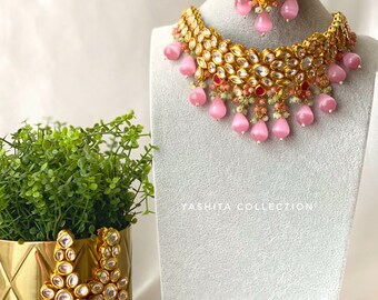 Puede incluir: Un conjunto de joyas de novia indias de color rosa y dorado con un collar, pendientes y un maang tikka. El collar presenta un diseño en capas con piedras rosas y transparentes, y los pendientes y el maang tikka tienen piedras rosas a juego.