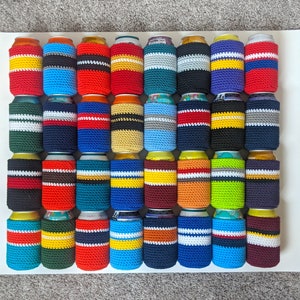 May include: A collection of 36 crocheted can coolers in various colors and patterns. The coolers are made with a variety of yarn colors, including red, blue, green, yellow, and white. Each cooler has a different pattern, including stripes, polka dots, and geometric designs.