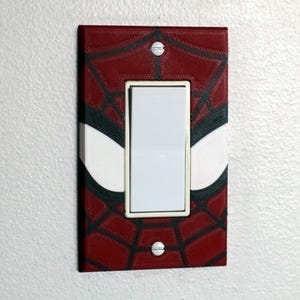 Puede incluir: Una placa de pared rectangular para interruptor de luz con un patrón de telaraña rojo y negro y grandes formas de ojos blancos. La cubierta está asegurada con dos tornillos plateados y rodea un interruptor basculante blanco.