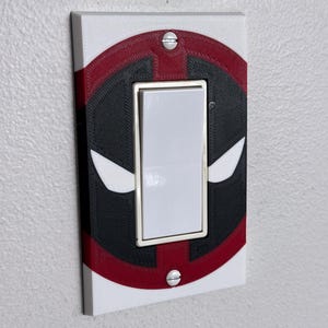 Puede incluir: Una cubierta de interruptor de luz blanca, roja y negra con un diseño inspirado en un personaje de cómic. La cubierta rectangular tiene un interruptor basculante central y está montada en una pared blanca.