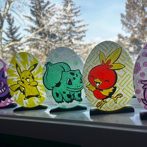 May include: Eight colorful egg-shaped ornaments featuring different Pokemon characters. The ornaments are displayed on a windowsill with a snowy landscape in the background.