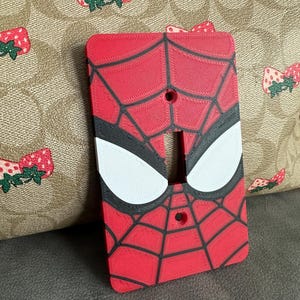 Pode incluir: Uma capa de interruptor de luz vermelha e preta com o tema Homem-Aranha. A capa apresenta um design de teia de aranha com formas de olhos brancos. O fundo é um tecido bege com um padrão de laço de morango.