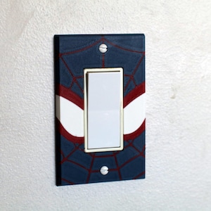 May include: A decorative light switch cover with a blue background and a red spiderweb pattern. The cover has white and red accents, and a white switch. It is mounted on a white wall.