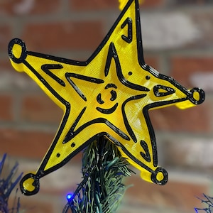 May include: A yellow star-shaped Christmas tree topper with a black outline and a smiley face. The star is made of plastic and has a shiny finish.