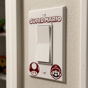 May include: A white rectangular light switch wall plate featuring the dark red text SUPER MARIO at the top. Below the central white rocker switch are two dark red graphic icons: a mushroom on the left and a Mario character face on the right.