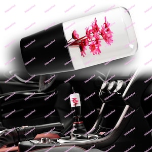 以下が含まれることがあります： ピンクの花が入った透明なトップが付いた車のギアシフトノブ。ノブは黒いベースと赤いアクセントが付いています。画像には、ダッシュボードやコンソールなど、車のインテリアも表示されています。