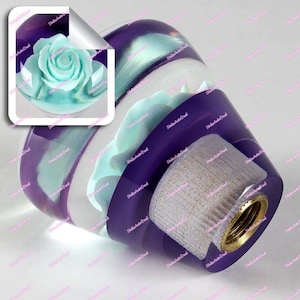 Op de afbeelding: Een decoratieve flesstopper met een lichtblauwe roos ingekapseld in heldere hars met paarse accenten. De stopper heeft een goudkleurige metalen basis en een getextureerde, lichtgekleurde inzet. Het ontwerp is artistiek en elegant.