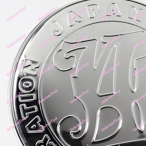 May include: A silver metal emblem with the words "JAPAN" and "ALL-MOTION" surrounding the number "67" in a stylized font.