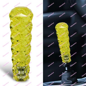 Puede incluir: Un pomo de palanca de cambios de coche de cristal amarillo facetado. El pomo tiene un diseño geométrico y una base metálica. El fondo es un interior de coche.