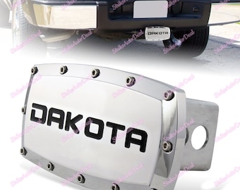 May include: Chrome hitch cover with a white background and black DAKOTA lettering. The cover has a rounded rectangular shape with a silver trim and multiple screws.
