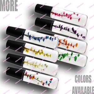 May include: An assortment of clear, rectangular lip gloss tubes with black caps, each containing dried flowers in various colors. The image includes the text "MORE" and "COLORS AVAILABLE".