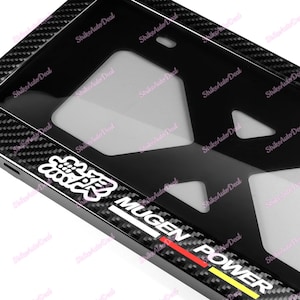 May include: Black carbon fiber license plate frame with a white "Mugen Power" logo and a red and yellow stripe. The frame has a black and white Japanese character logo.