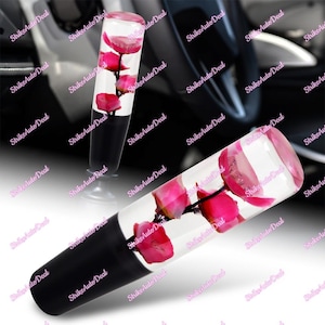 May include: A car gear shift knob with a clear, cylindrical design. The knob is filled with pink flowers and black accents. The text "Strike Auto Deal" is repeated on the background.