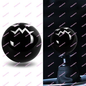 May include: A black shift knob featuring a white zigzag design. The spherical knob has a glossy finish. The image presents the knob in two contexts: isolated and fitted to a gear stick.