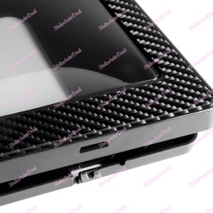 May include: A black plastic car part with a carbon fibre pattern. The part has a rectangular shape with a small opening on the side.