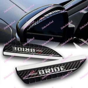 Könnte beinhalten: Zwei schwarze Carbon-Spiegelabdeckungen für Autos mit einem weißen Streifen und dem Wort "BRIDE" in weißen Buchstaben.