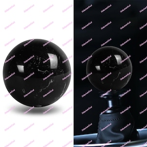 May include: A black, spherical gear knob with white marbling. The knob is shown in two views, one isolated and the other attached to a black leather boot. Suitable for automotive use.