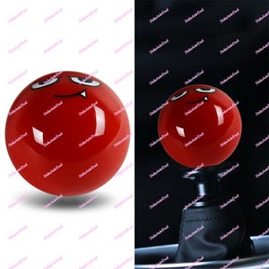 May include: A red shift knob with a cartoon devil face. The shift knob is spherical with white eyes, a curved smile, and two small fangs. The image shows the shift knob on a black gear shift.