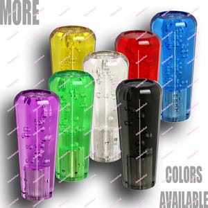 May include: A collection of seven translucent gear shift knobs in various colors: purple, yellow, green, clear, red, black, and blue. Each knob has a bubble-like design inside. The words "MORE" and "COLORS AVAILABLE" are in the image.