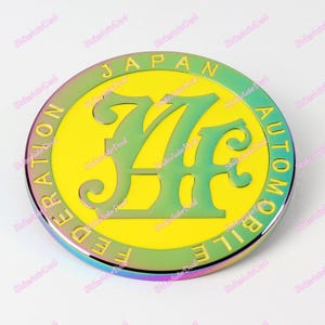 May include: A yellow and green metal emblem with the text "JAPAN AUTOMOBILE FEDERATION" surrounding a stylized "7" in the centre. The emblem has a rainbow iridescent finish.
