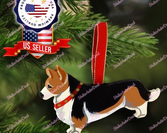 May include: Wooden corgi dog Christmas ornament with a red ribbon. The ornament features black, white, brown, and red paint details. The ornament is suspended in front of a green Christmas tree.