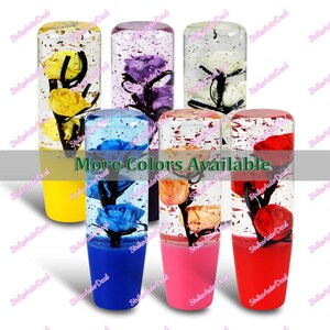 May include: A collection of colorful, decorative car shift knobs. Each knob features a preserved rose in a clear, cylindrical casing filled with glitter. The base of each knob is a solid color, with options including yellow, purple, white, blue, orange, pink, and red.