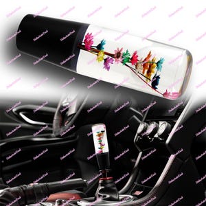 Può includere: Un pomello del cambio trasparente con una parte superiore nera, riempito di fiori colorati. I fiori includono fiori rosa, gialli e blu. Il pomello è cilindrico e sembra essere fatto di vetro o plastica trasparente.