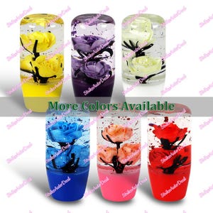 May include: A collection of colorful car shift knobs with embedded flowers. The knobs are in various colors, including yellow, purple, white, blue, peach, and red. The text "More Colors Available" is displayed across the image.