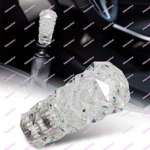 Könnte beinhalten: Ein klarer, facettierter Kristall-Schaltknauf. Der Knauf hat oben ein Totenkopf-Design und ist als Ersatz für einen Standardschaltknauf konzipiert. Das Bild zeigt den Knauf in einem Autoinnenraum.