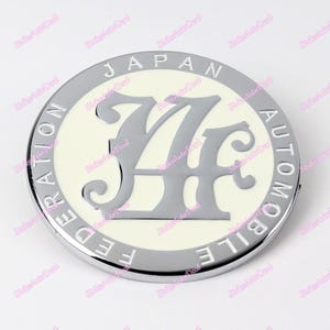 Op de afbeelding: Een zilveren en wit embleem met de tekst "JAPAN AUTOMOBILE FEDERATION" rond een gestileerd "JF"-logo.