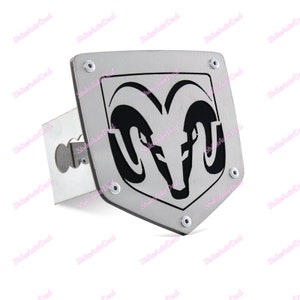 Pode incluir: Uma cobertura de engate de reboque prateada com o logotipo Dodge Ram em preto. O logotipo é uma cabeça de carneiro dentro de uma moldura em forma de escudo. A cobertura é feita de metal e tem uma base retangular com um orifício para o engate.