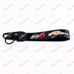 May include: Black key chain with a white car and red and white logo that says "Initial D" in Japanese. The key chain has a metal ring.