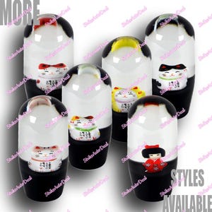 以下が含まれることがあります： 黒、白、赤を基調とした、様々なデザインのシフトノブのコレクション。 招き猫や日本人形のモチーフが特徴です。「STYLES AVAILABLE」の文字が見えます。