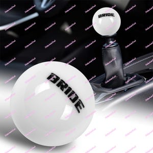 May include: White shift knob with the word "BRIDE" in black lettering. The shift knob is round and glossy, with a black base and a leather boot. The image shows the shift knob installed in a car.