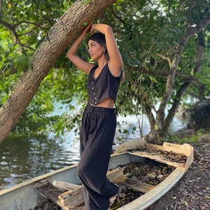 Puede incluir: Una mujer con un top negro sin mangas y pantalones negros anchos se encuentra de pie en un bote de madera. El bote está en un camino de tierra junto a una masa de agua. La mujer se apoya en un tronco de árbol grande.