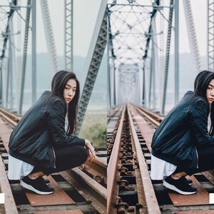 May include: A woman wearing a black jacket and black pants sits on a set of railroad tracks on a bridge. The bridge is made of metal and has a gray background.