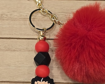 Puede incluir: Un llavero dorado con un pompón rojo y esponjoso y un dije con una mujer de dibujos animados. El dije está unido a una cadena de cuentas rojas y negras. El llavero tiene un cierre de mosquetón.