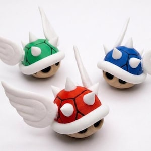 May include: Three colorful, winged Koopa shells in green, blue, and red. Each shell has white spikes, a white rim, and white wings. The shells are set against a white background.