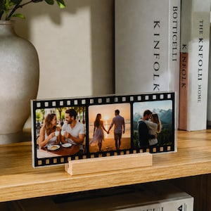 May include: A rectangular acrylic photo frame designed to resemble a strip of 35mm film, featuring three distinct color photographs of a couple. The clear frame is supported by a light-colored wooden base, displaying images of shared moments.