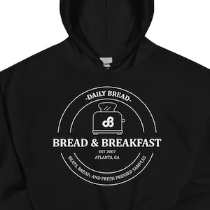 Könnte beinhalten: Schwarzer Kapuzenpullover mit einem weißen, kreisförmigen Logo, das "DAILY BREAD" und "BREAD & BREAKFAST" mit einer Toaster-Grafik zeigt. Der Text "EST 2007 ATLANTA, GA" ist ebenfalls enthalten. Der untere Teil des Logos lautet "BEATS BREAD AND FRESH PRESSED SAMPLES".
