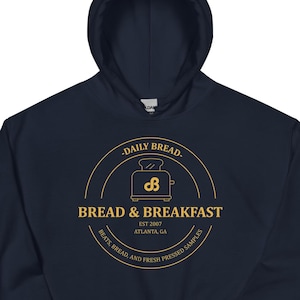 Può includere: Felpa con cappuccio blu navy con un logo circolare dorato che recita "DAILY BREAD" e "BREAD & BREAKFAST" con una grafica di tostapane. È incluso anche il testo "EST 2007 ATLANTA, GA".
