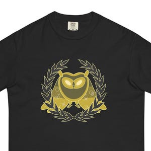 May include: A black short-sleeve t-shirt featuring a centered graphic of a stylized owl in shades of yellow and gold. The owl is framed by a circular laurel wreath design in a light yellow outline, with small star and circle accents on its wings.