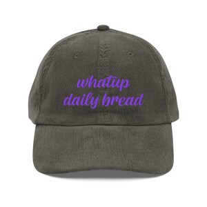 Op de afbeelding: Een donkergrijze corduroy baseballpet met de geborduurde tekst whatup daily bread in een felpaars cursief lettertype op het voorpaneel. De pet heeft een gestructureerde kroon, een gebogen klep en bijpassende stiksels over het gehele ontwerp.