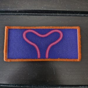 May include: A rectangular patch with a blue background and a pink and white heart-shaped design. The patch has an orange border and is likely made of fabric.