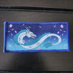 May include: A rectangular patch with a white and teal dragon design on a blue gradient background. Pink floral elements and a blue border frame the dragon. The patch is a decorative item.